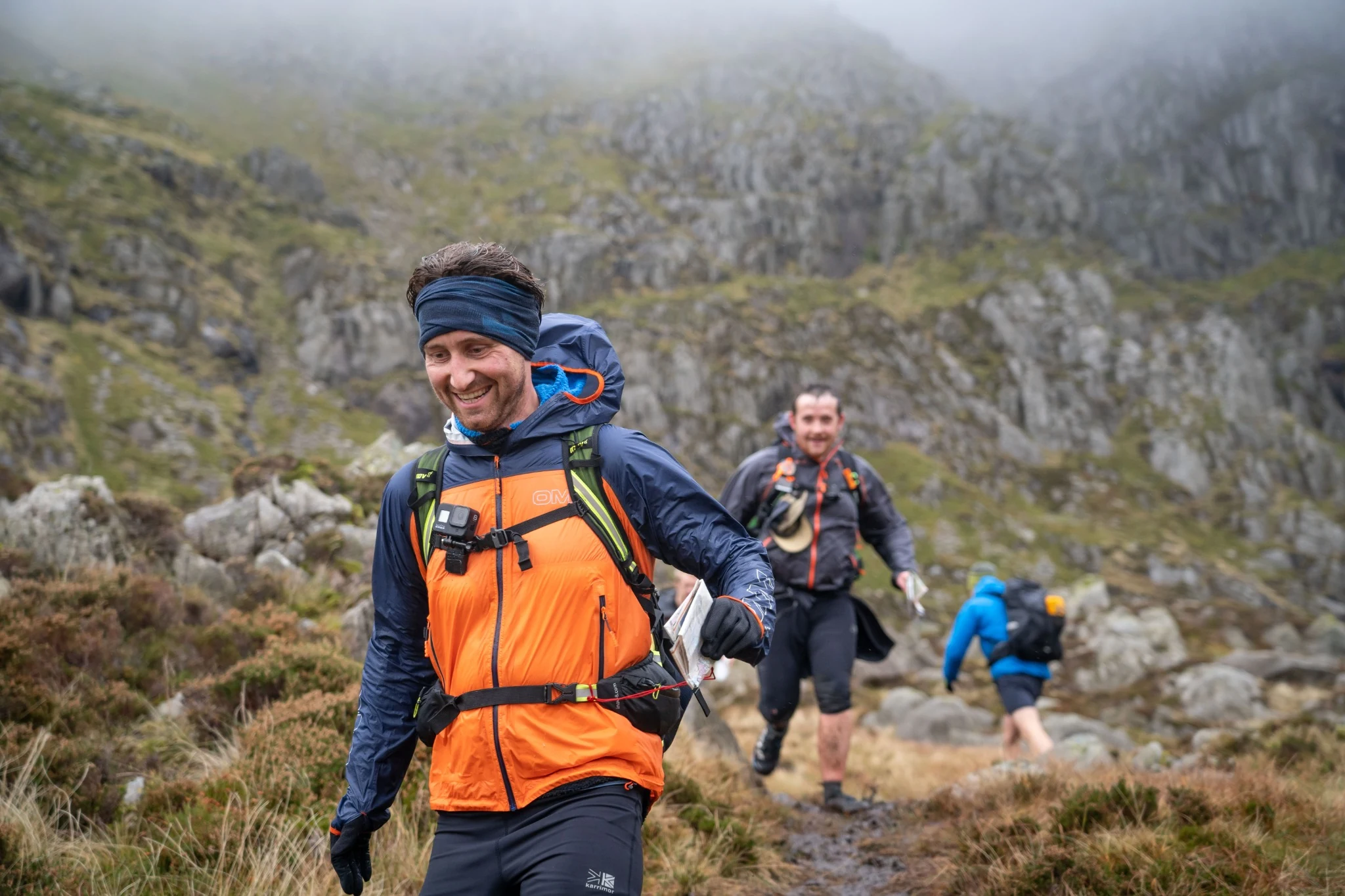 Two OMM competitors carrying packs through open grassland beneath a blue sky