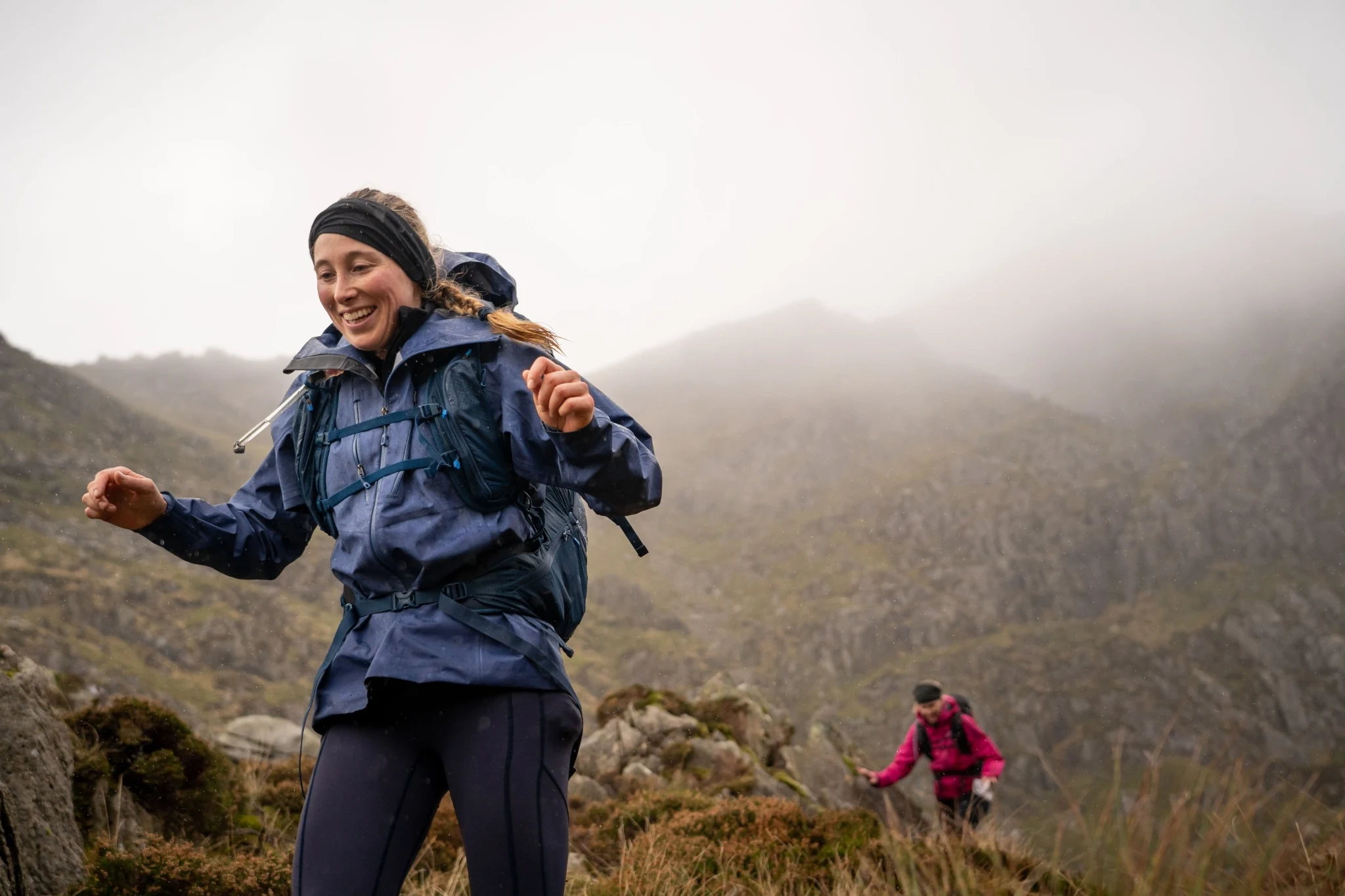 A smiling OMM competitor carrying a pack across open mountain terrain